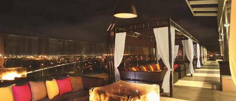 A rooftop lounge with a fire feature, seating, and a view of the cityscape.