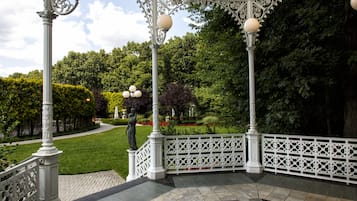 Un belvédère agrémenté de balustrades et de colonnes blanches décorées, entouré d’un jardin bien entretenu, avec des haies et des arbres.