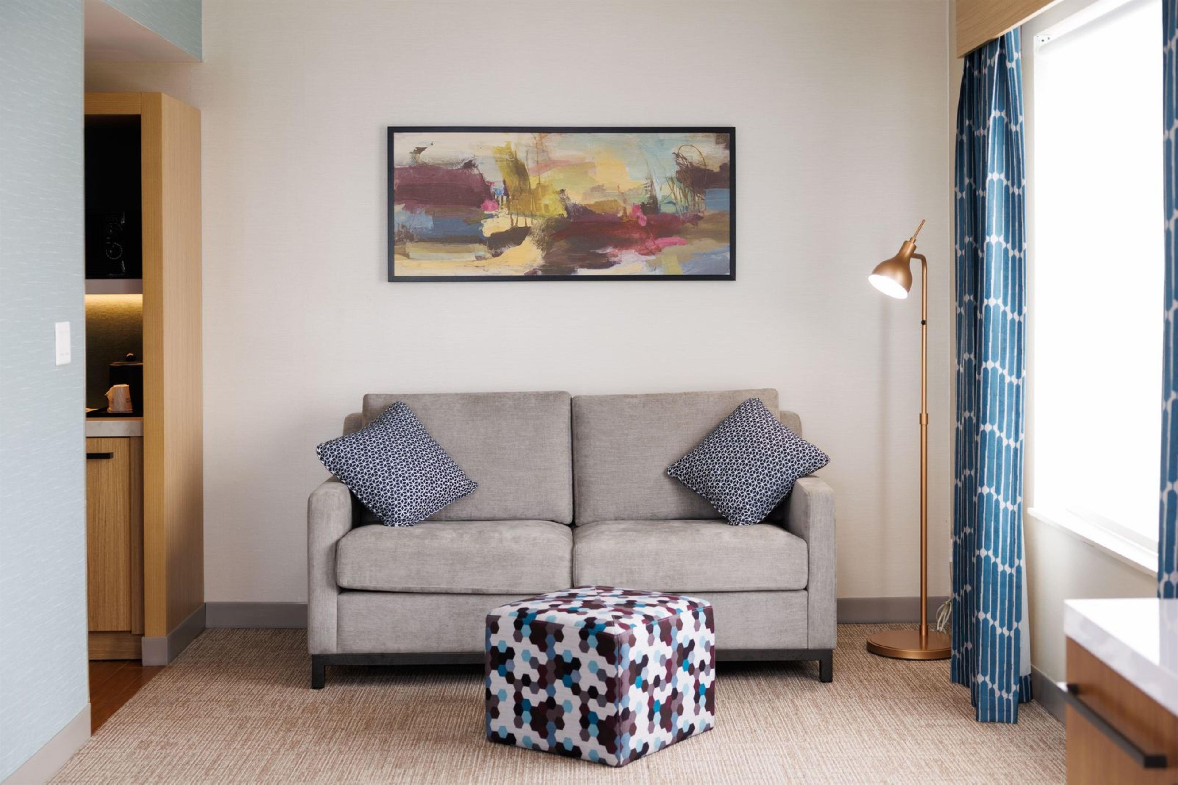 A living room with a grey sofa, patterned cushions, a colorful abstract painting, a floor lamp, and a patterned ottoman.