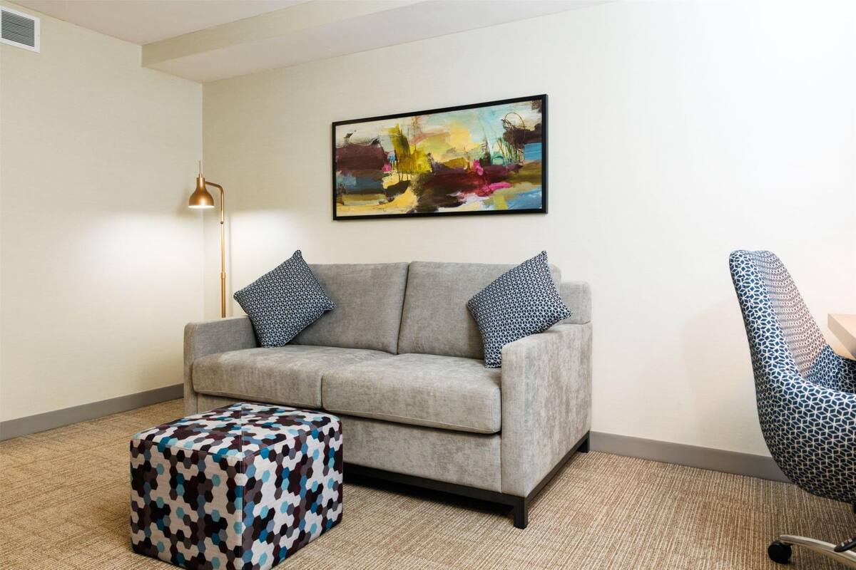 Un salón con un sofá gris, un taburete estampado y una silla azul con diseño. Una lámpara de pie y un cuadro abstracto de colores adornan la pared.