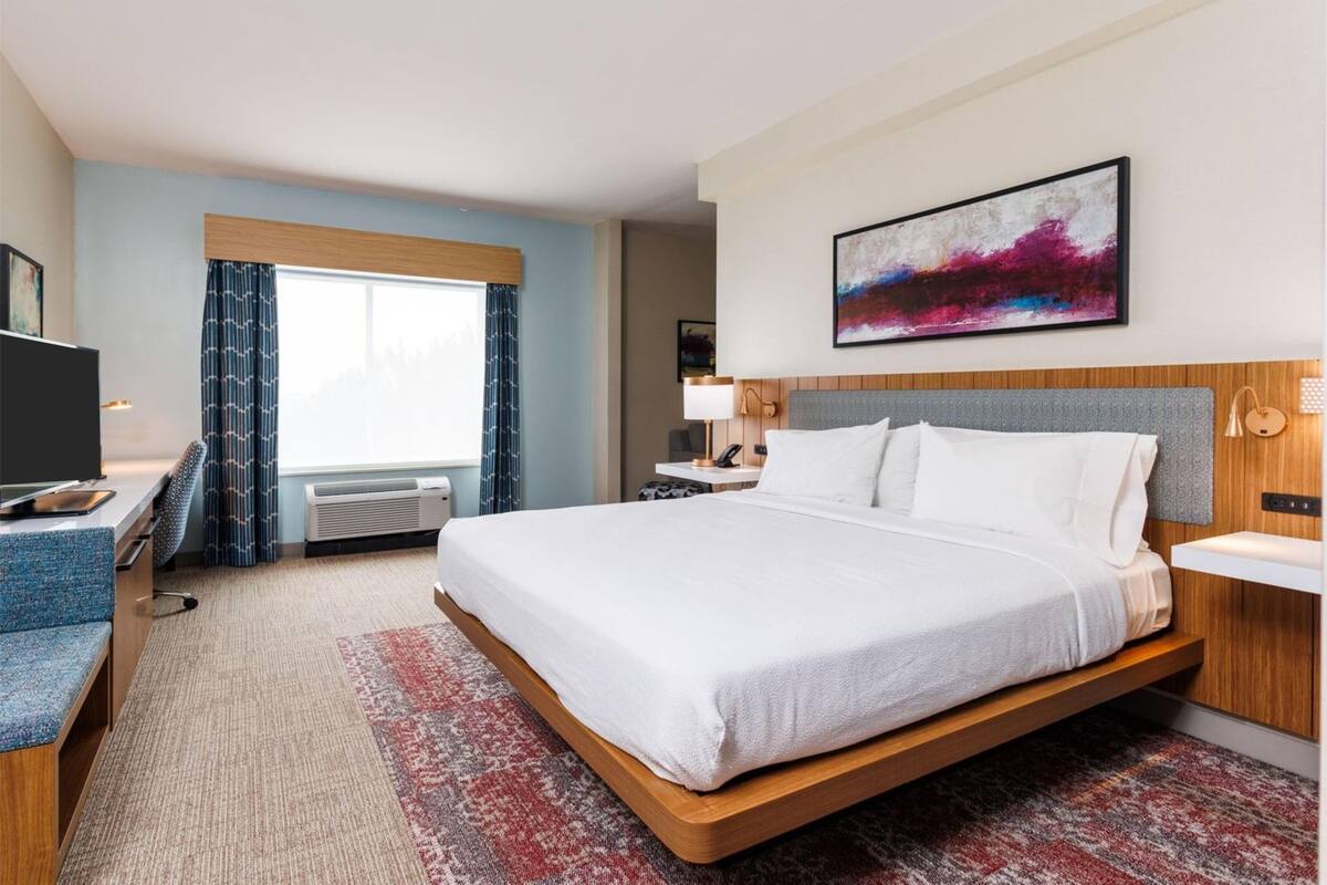 Habitación de hotel con cama, escritorio, silla, televisor, ventana con cortinas, un cuadro en la pared y mesita de noche con lámpara.