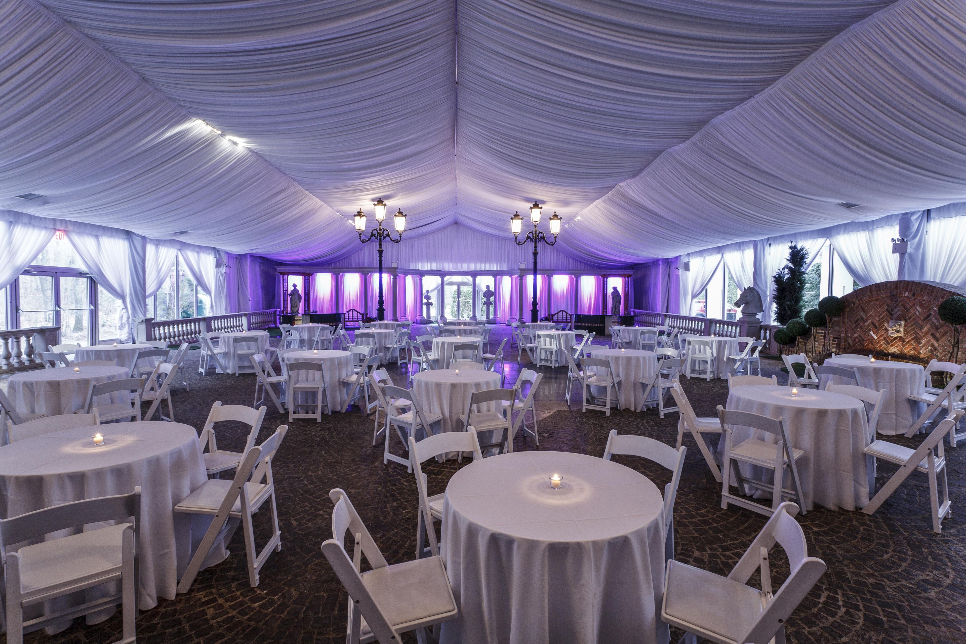 A large tented event space with round tables and white chairs, chandeliers, and a brick wall with a horse statue.