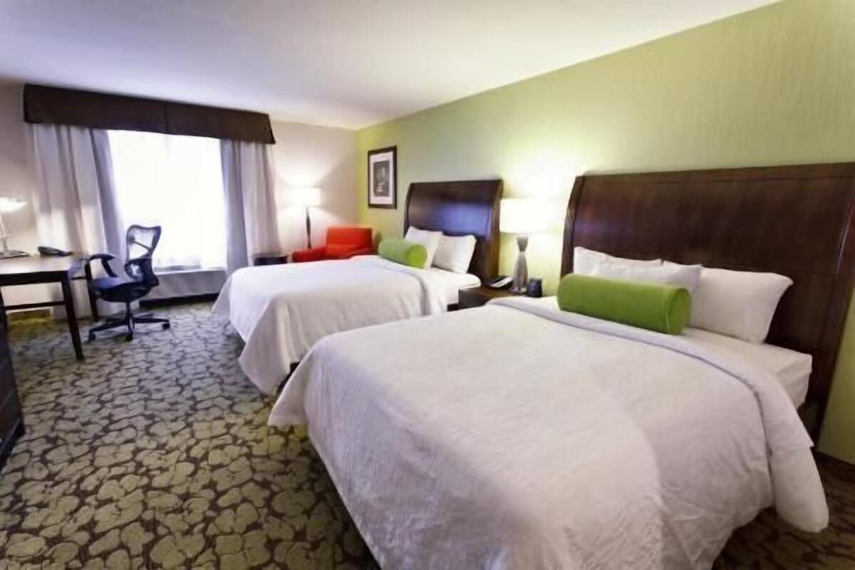 Habitación de hotel con dos camas, un escritorio, una silla y un ventanal con cortinas.