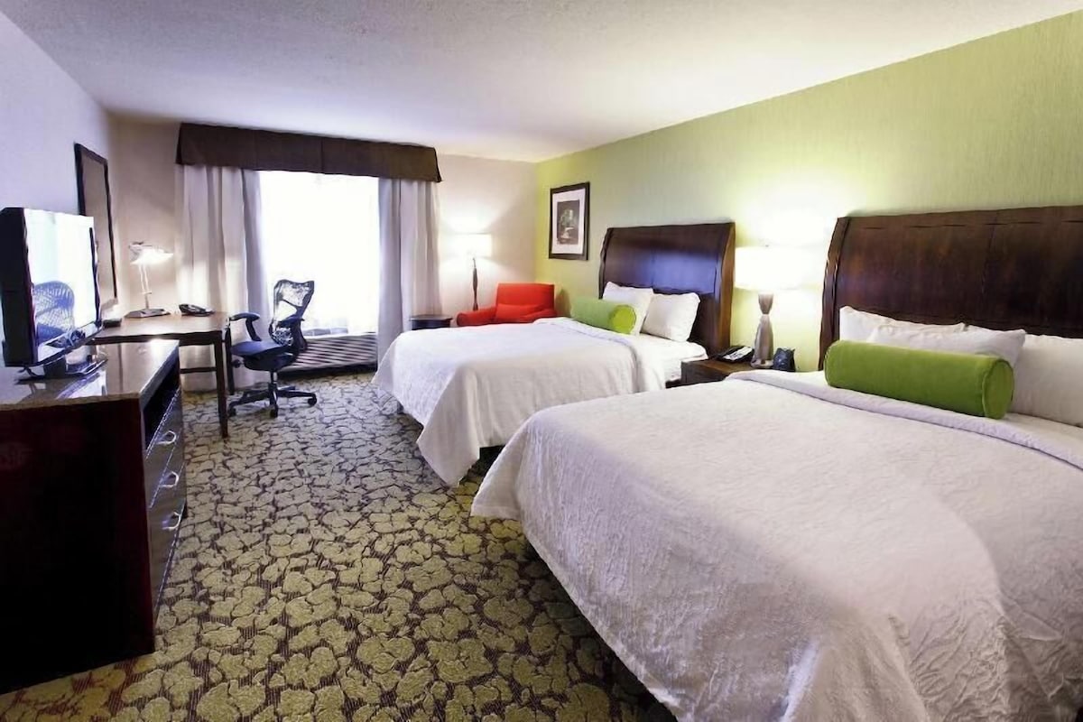 A hotel room with two beds, a desk with a chair, a lamp, and a window with curtains.
