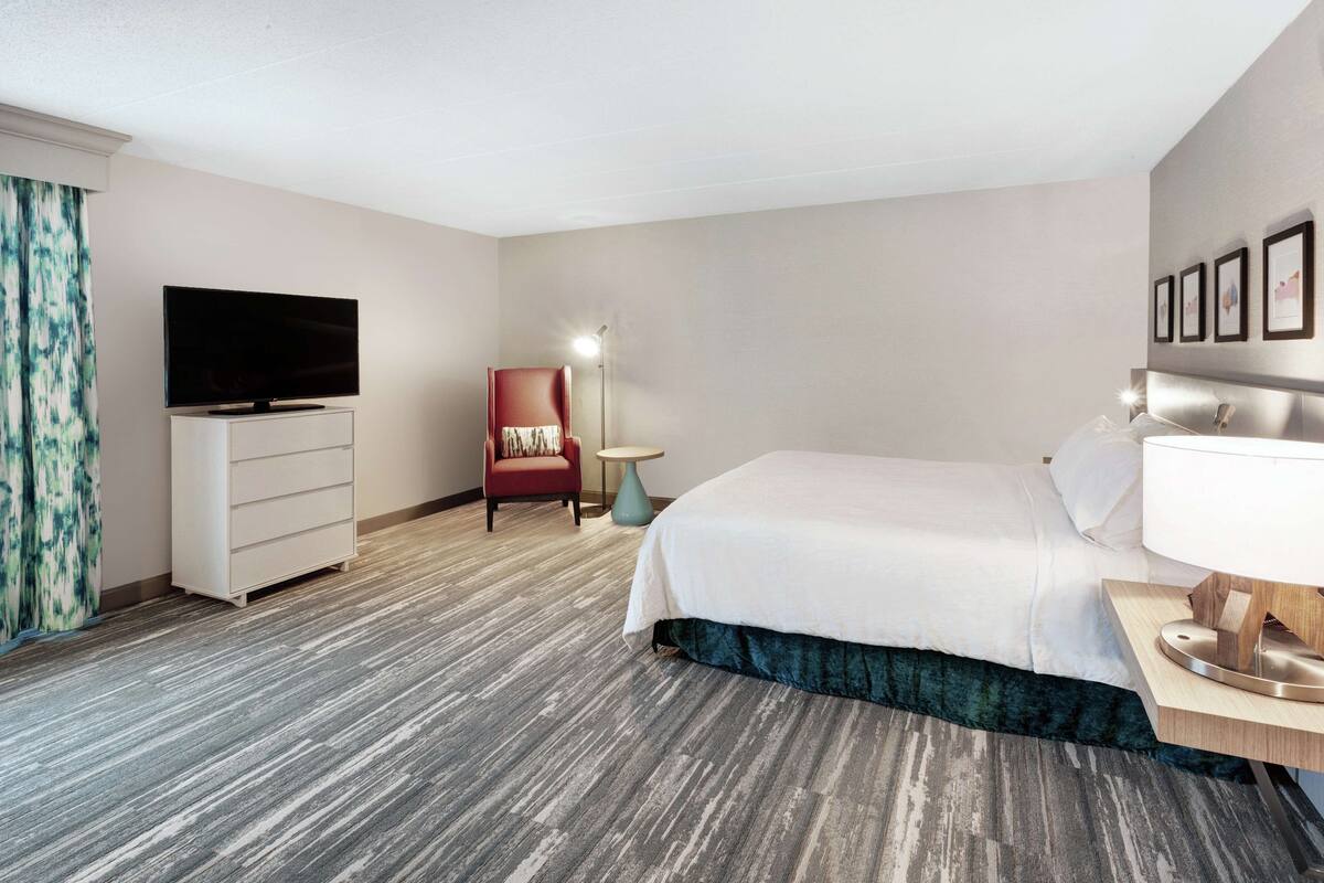A hotel room with a bed, a TV, a red chair, a small table, and a lamp.
