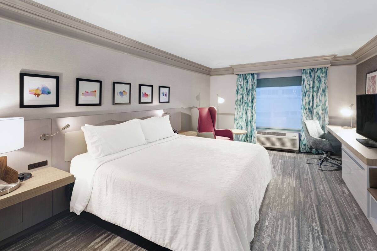 A hotel room with a large bed, a desk with a computer, a chair, and a TV. There are framed pictures on the wall and a window with curtains.