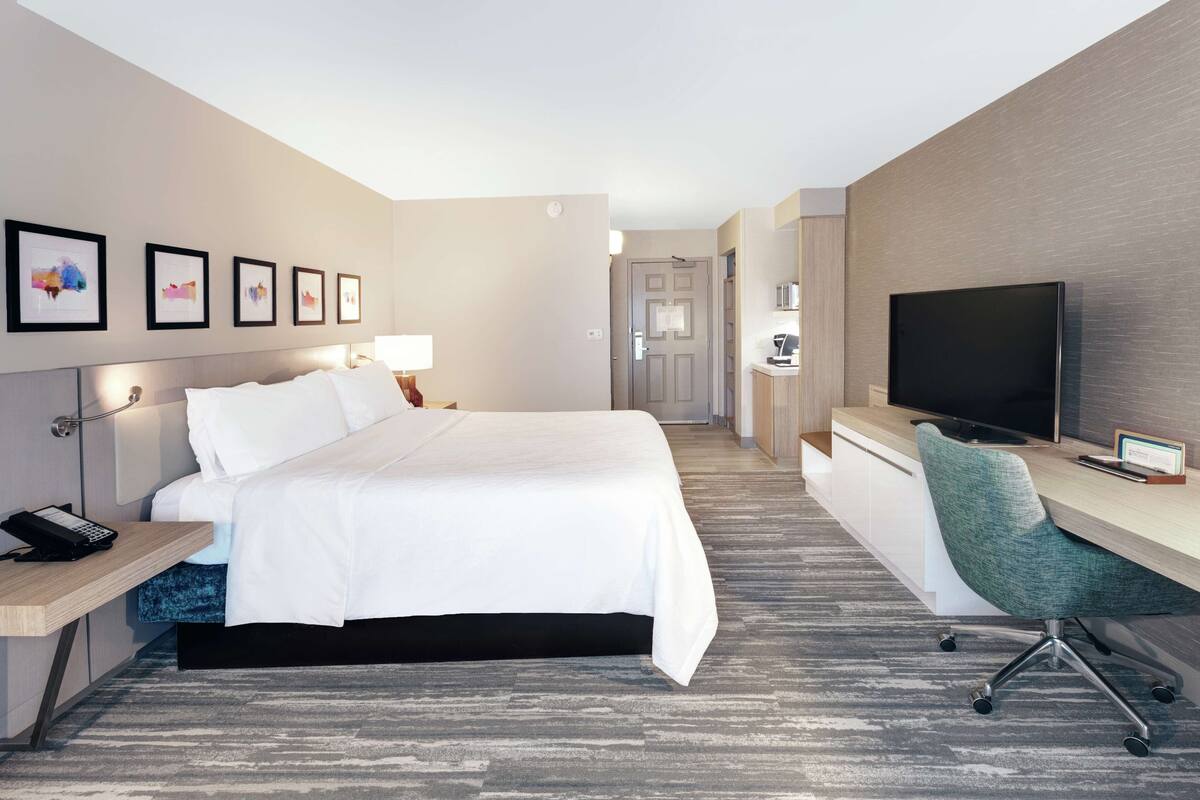 A modern hotel room with a neatly made bed, a desk with a computer, a TV, and framed pictures on the wall.