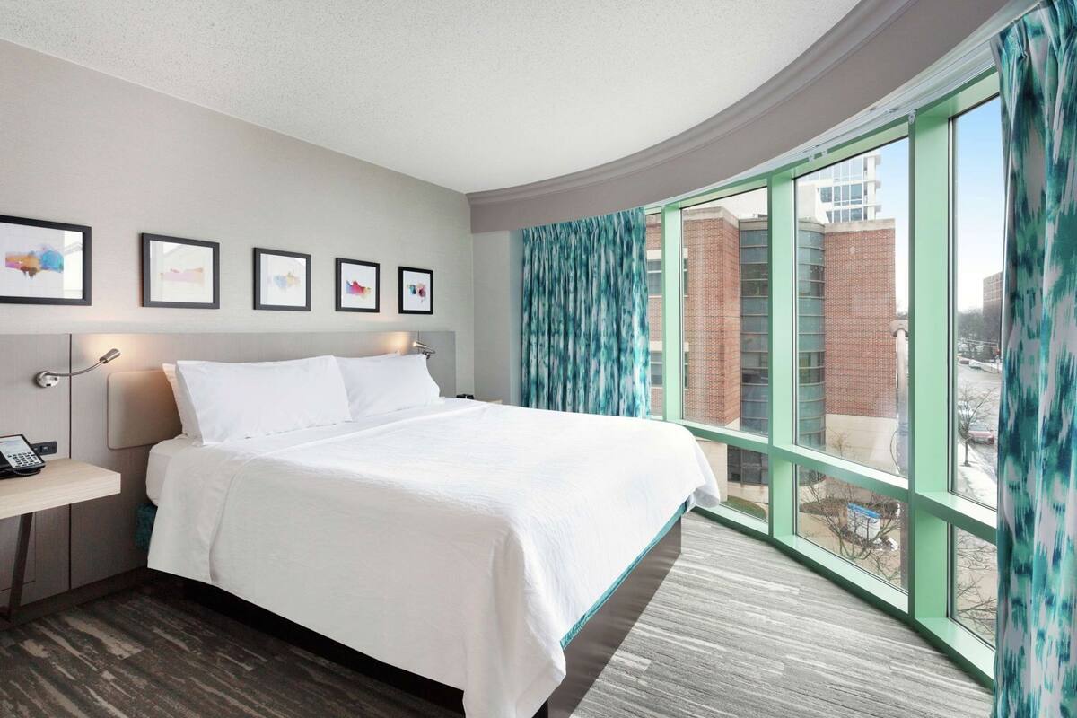 A hotel room with a large bed, a bedside table with a phone, a wall-mounted lamp, and a window with curtains.