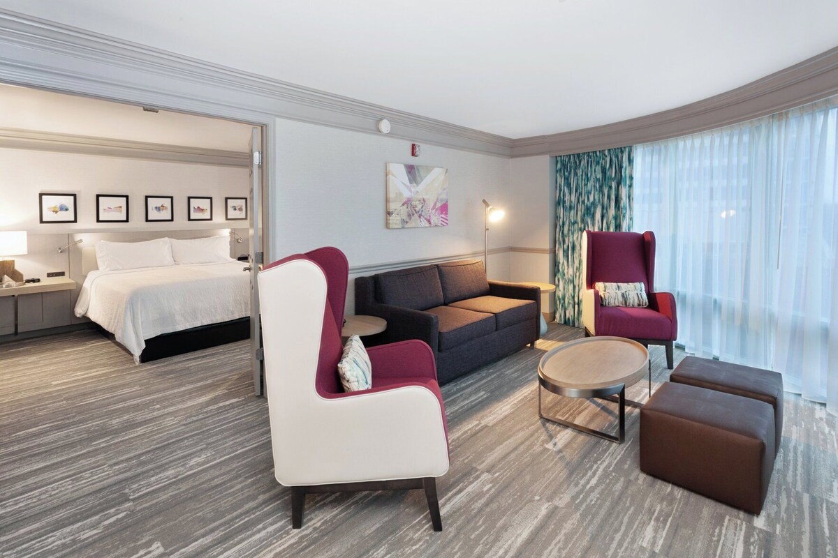 A hotel room with a bed, a sofa, a chair, a small table, and a large window.