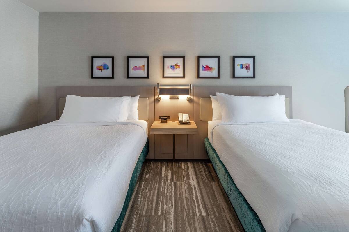 A hotel room with two beds, a nightstand with a phone and lamp, and four framed pictures on the wall.