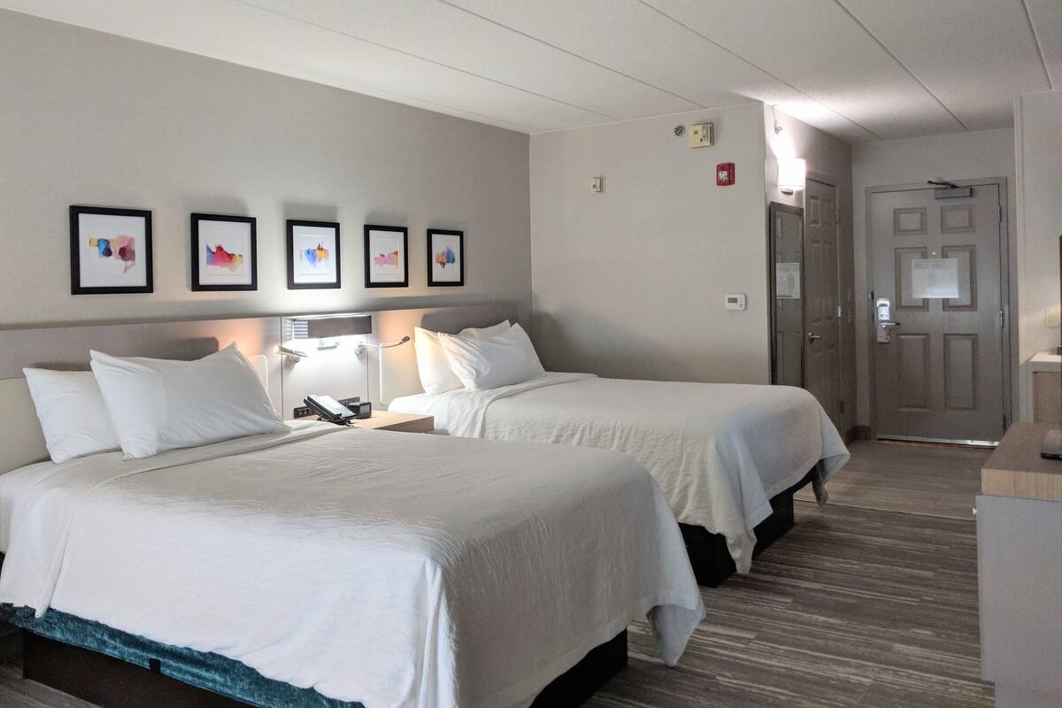 A hotel room with two beds, a desk, a chair, a lamp, and framed artwork on the wall.
