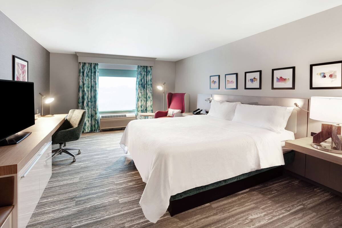 A hotel room with a bed, desk, chair, TV, and artwork on the wall.