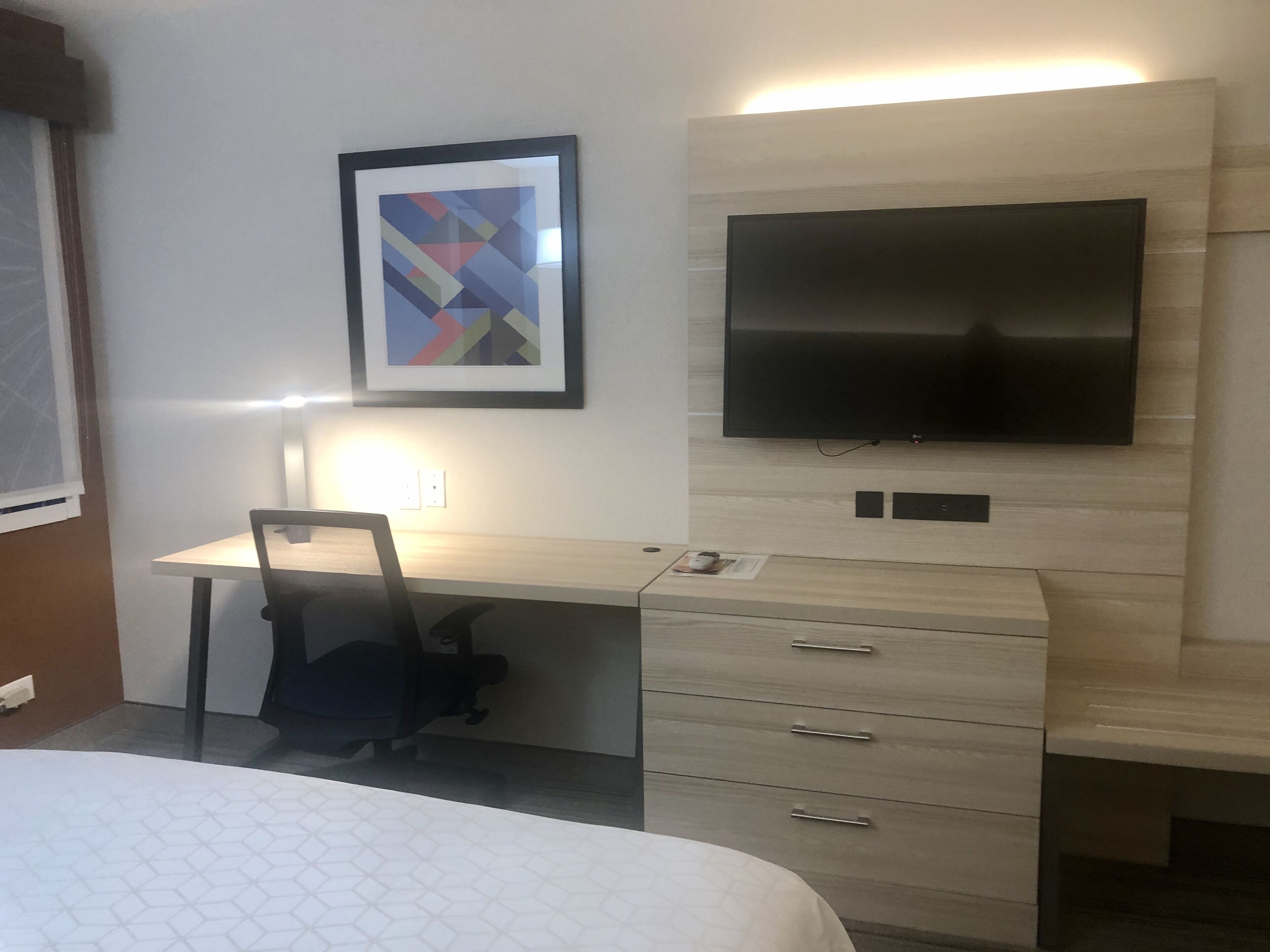 A hotel room with a bed, a desk, a chair, a flat-screen TV mounted on the wall, and a framed abstract artwork above the desk.