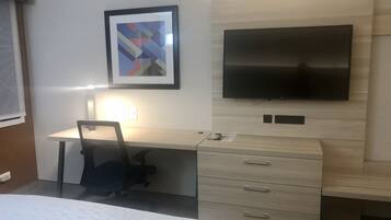 A hotel room with a bed, a desk, a chair, a flat-screen TV mounted on the wall, and a framed abstract artwork above the desk.