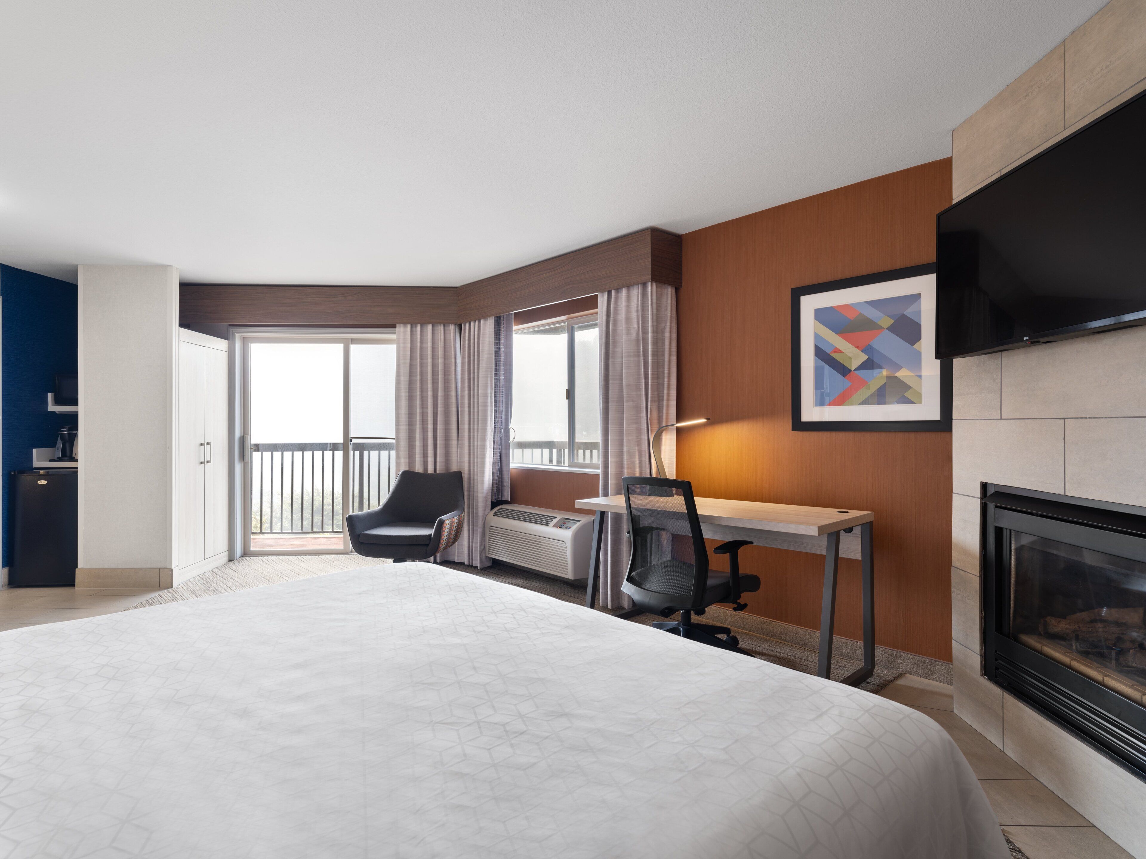 A hotel room with a large bed, a desk, a chair, a fireplace, and a TV.
