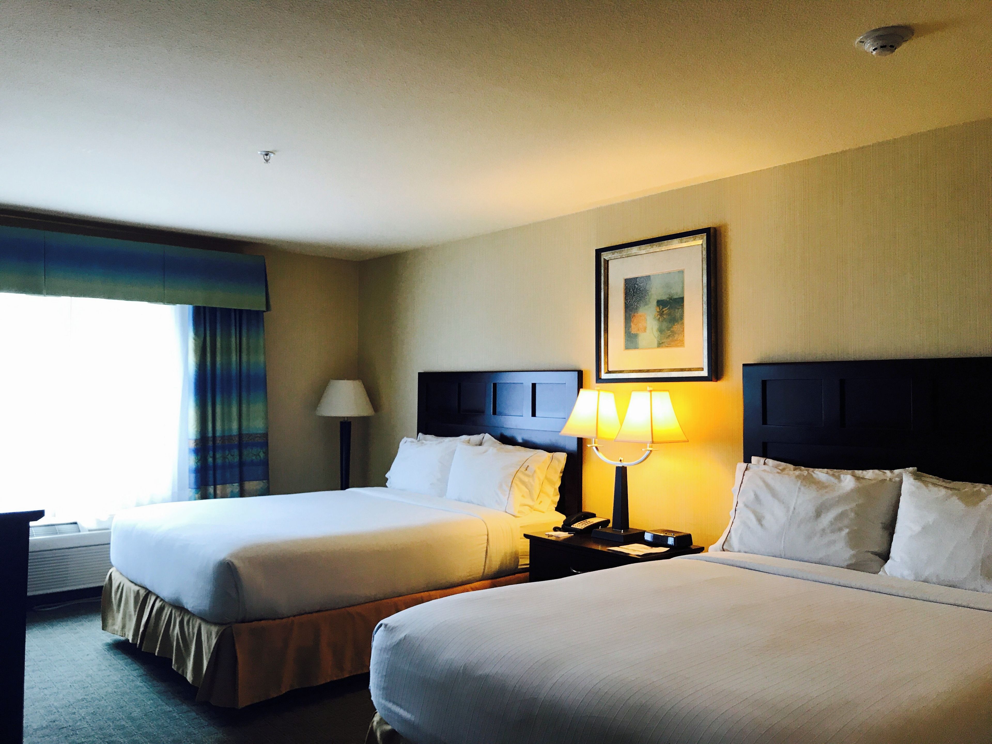 A hotel room with two beds, a nightstand with a lamp, a painting on the wall, and a window with curtains.