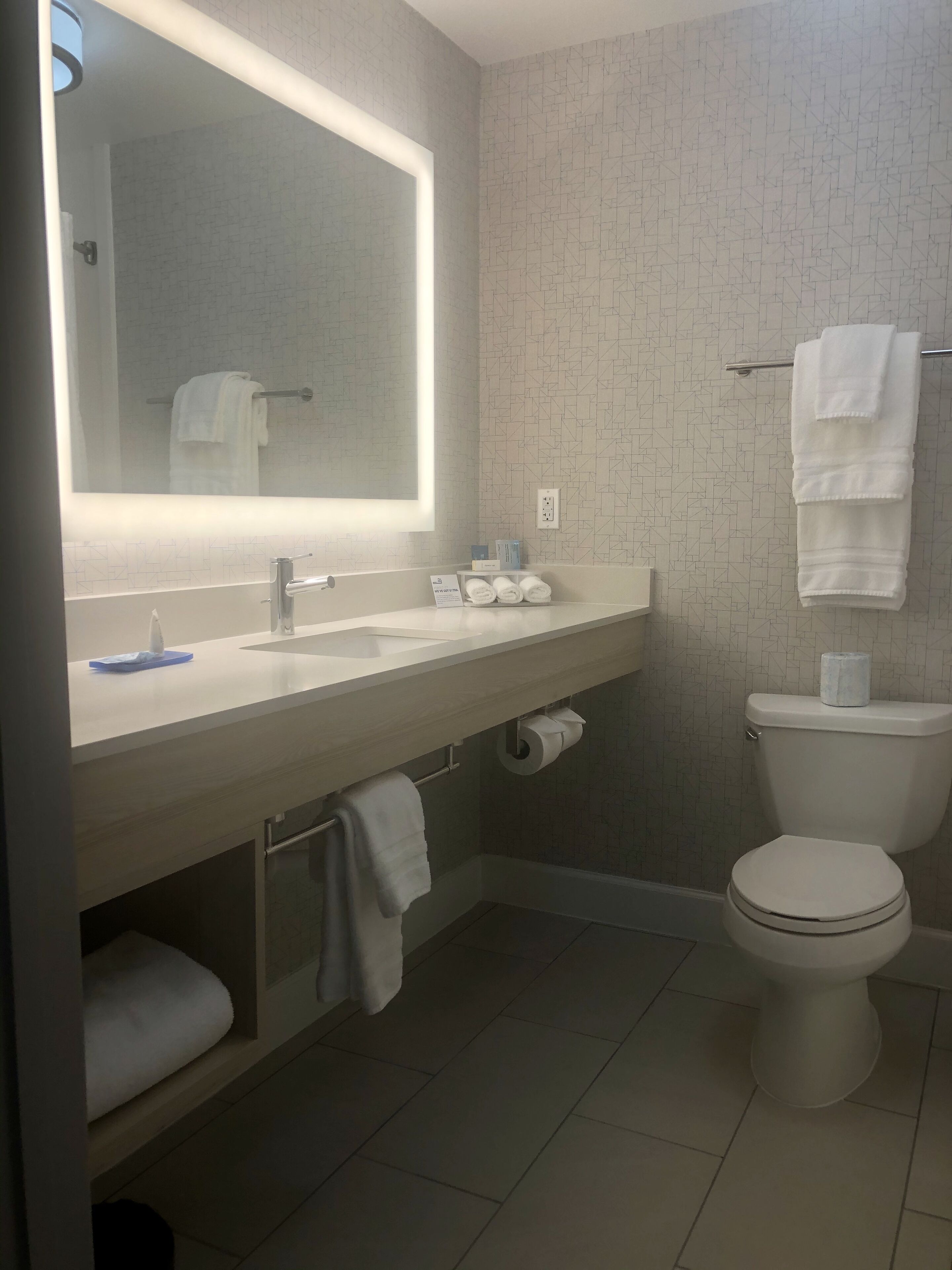A modern bathroom with a white sink, mirror, and toilet. There are towels hanging on the wall and a shelf with toiletries.
