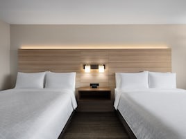 Habitación de hotel con dos camas, cabecera de madera, mesita de noche con un reloj y lámpara de pared.