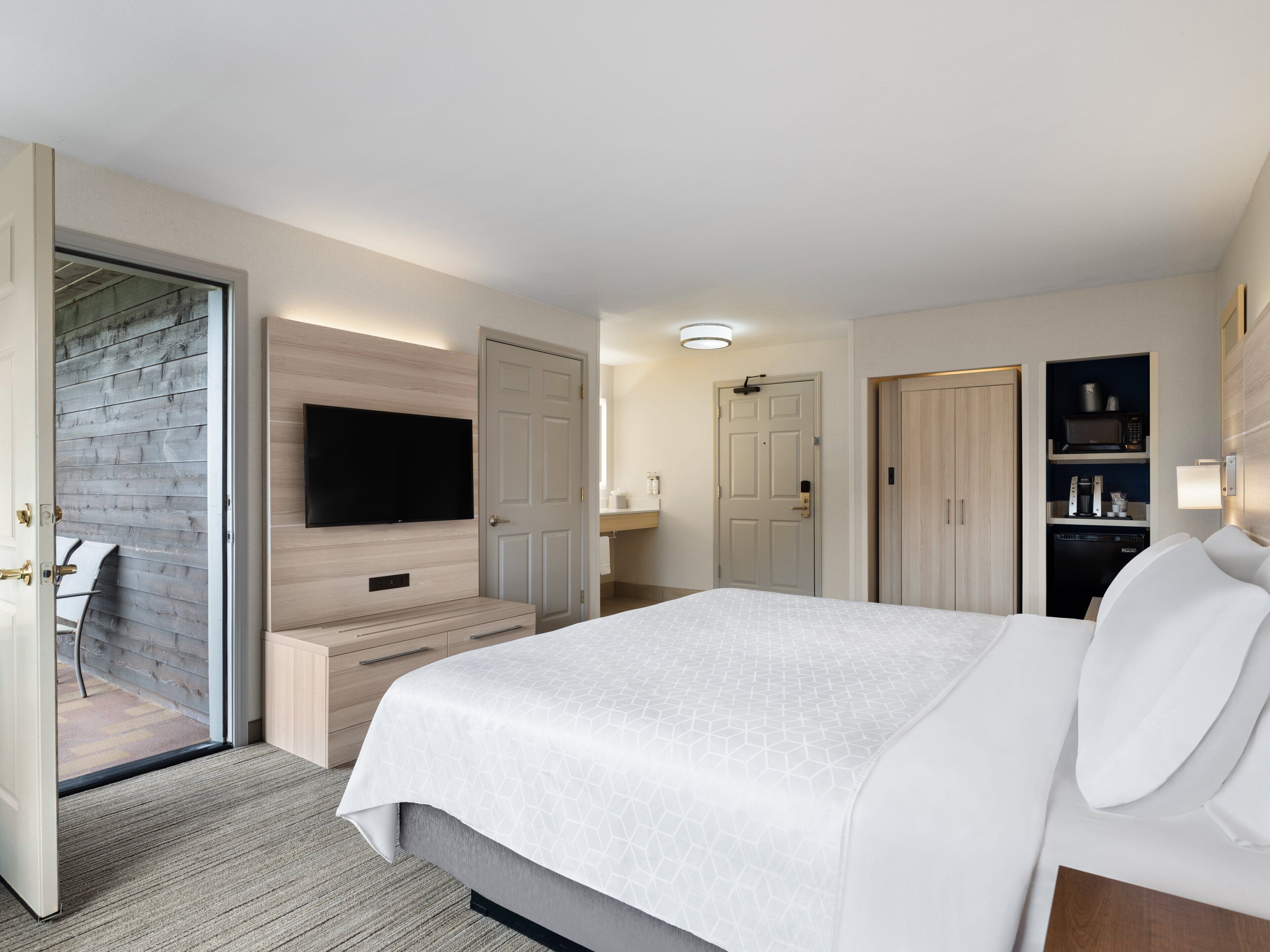 A modern hotel room with a large bed, a flat-screen TV mounted on the wall, a wooden bench, a door leading to a balcony, and a built-in shelf with a microwave.