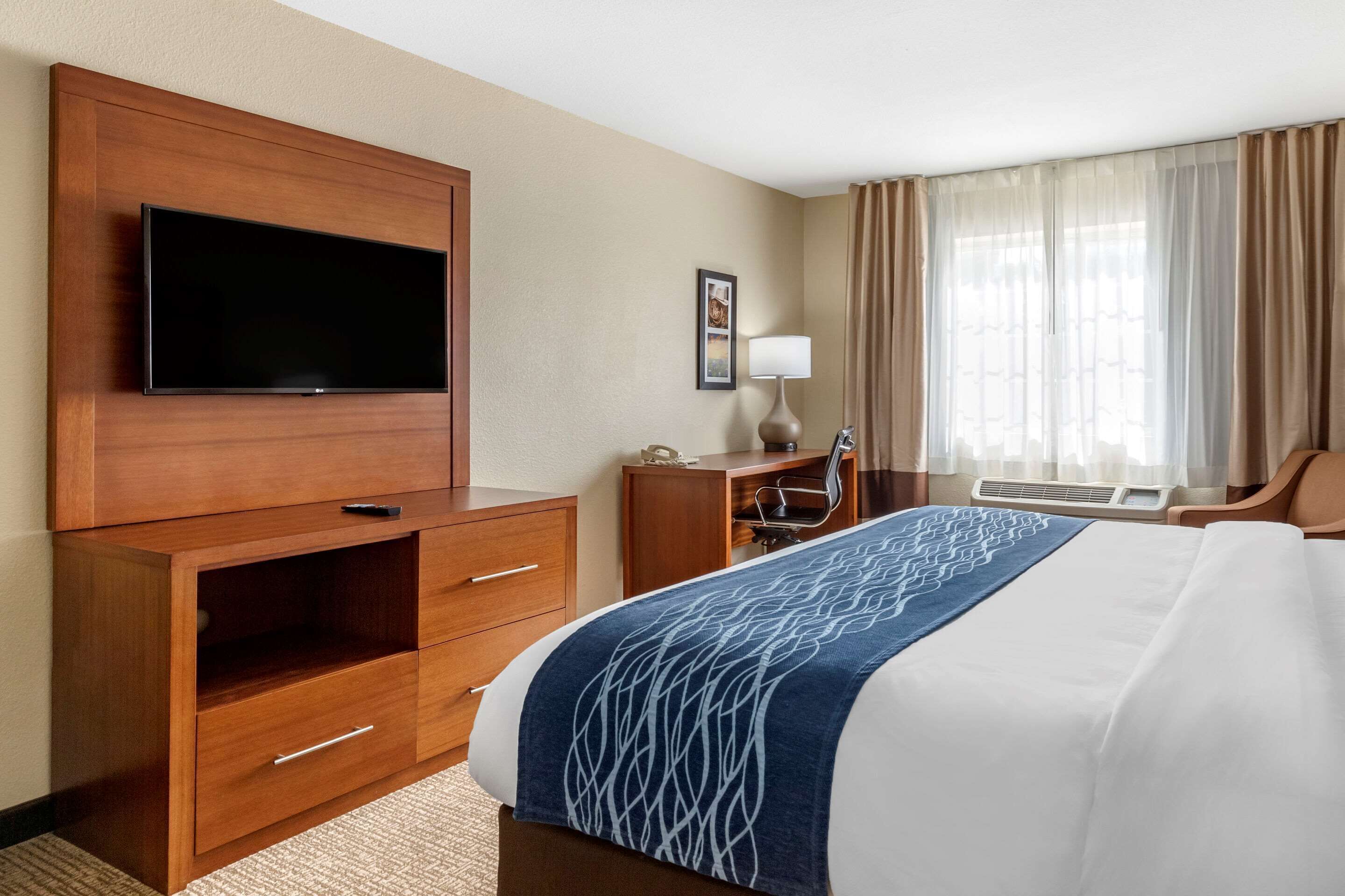 A hotel room with a large bed, a flat-screen TV mounted on a wooden cabinet, a desk with a chair, and a window with curtains.