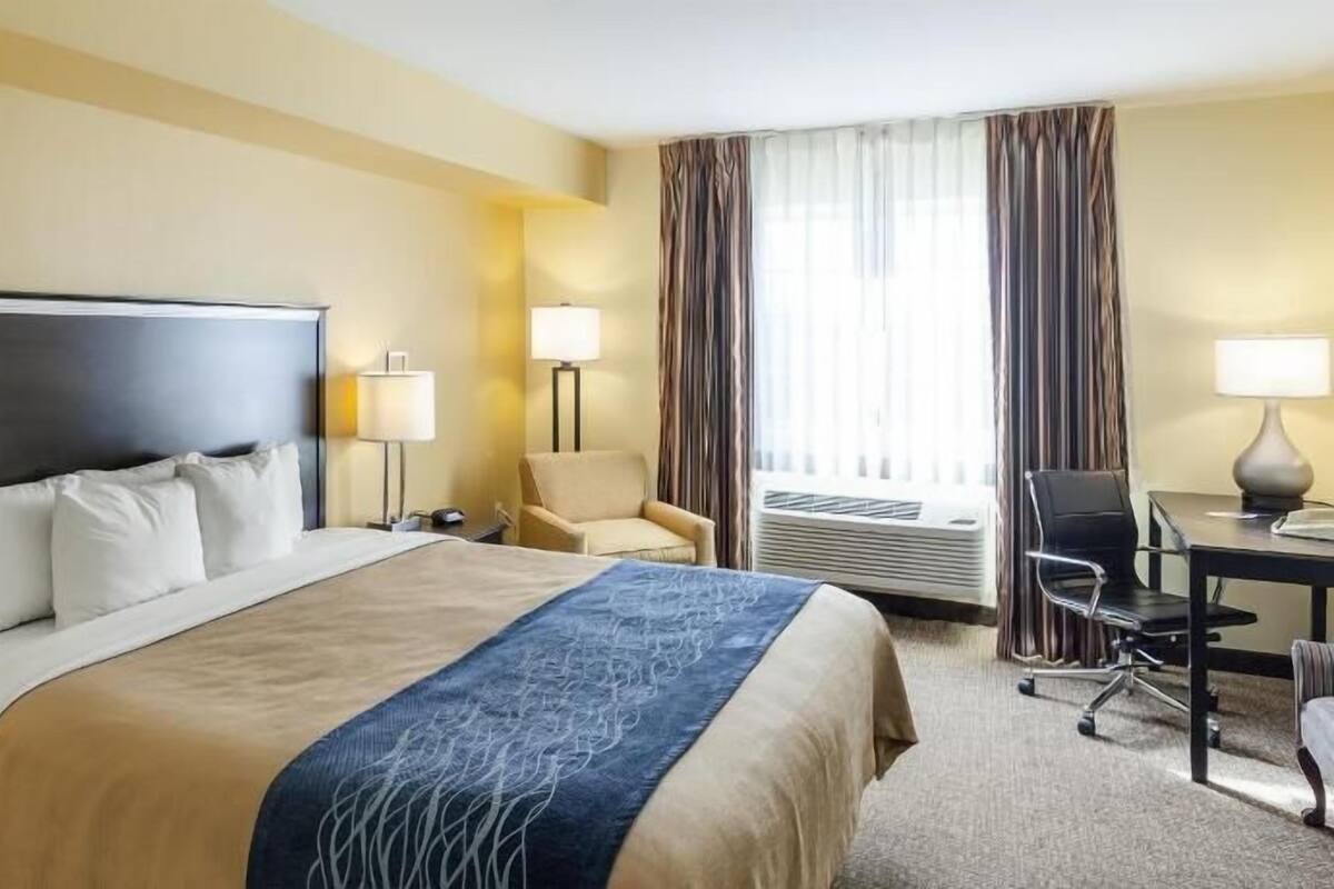 Habitación de hotel con una cama grande, dos mesitas de noche, un escritorio con una silla, otra silla y una ventana con cortinas.