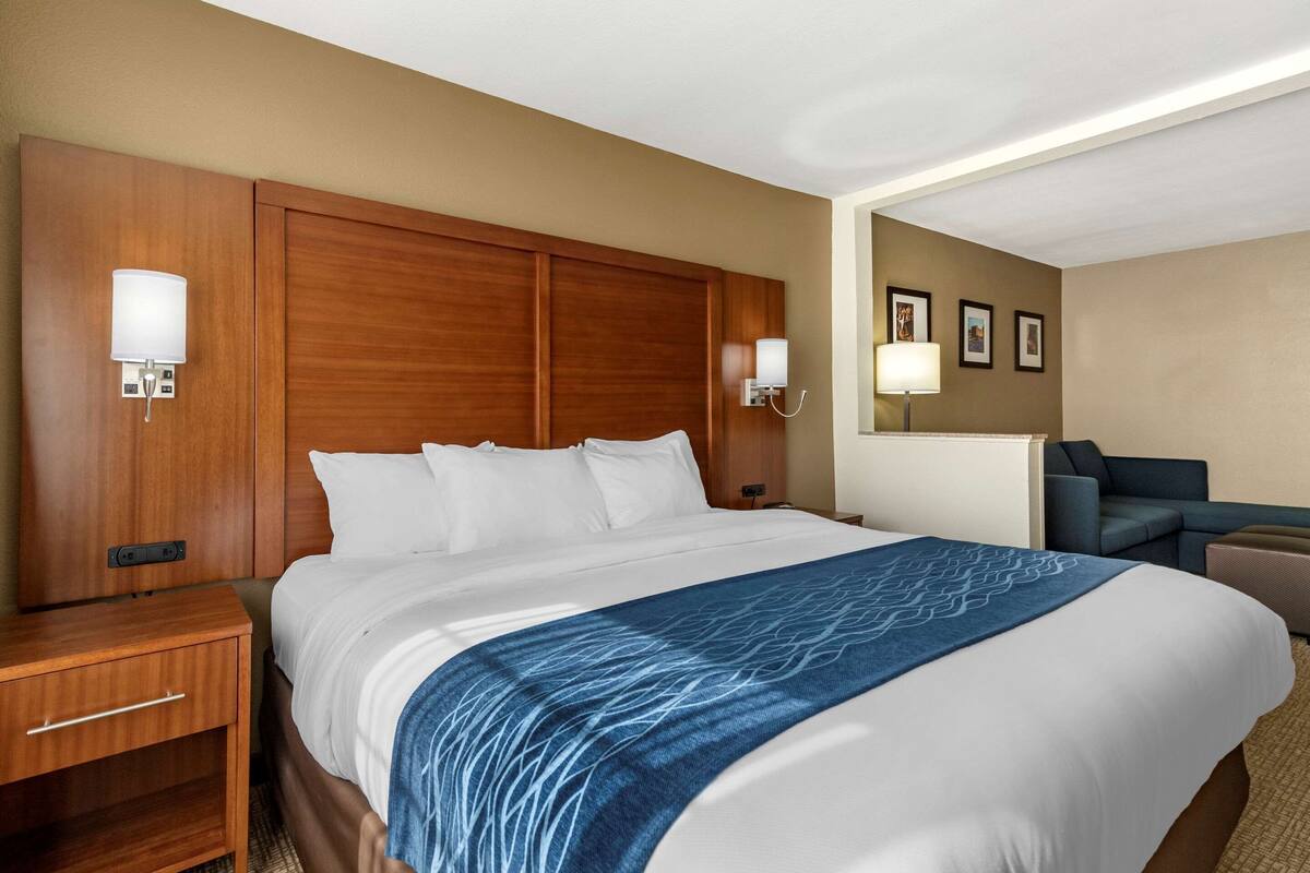 Habitación de hotel con una cama grande, cabecera de madera, dos mesitas de noche y una zona de estar con una silla y reposapiés.