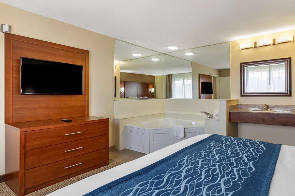 Habitación de hotel con cómoda de madera, televisor de pantalla plana grande, bañera, espejo grande y baño con lavamanos y tocador.