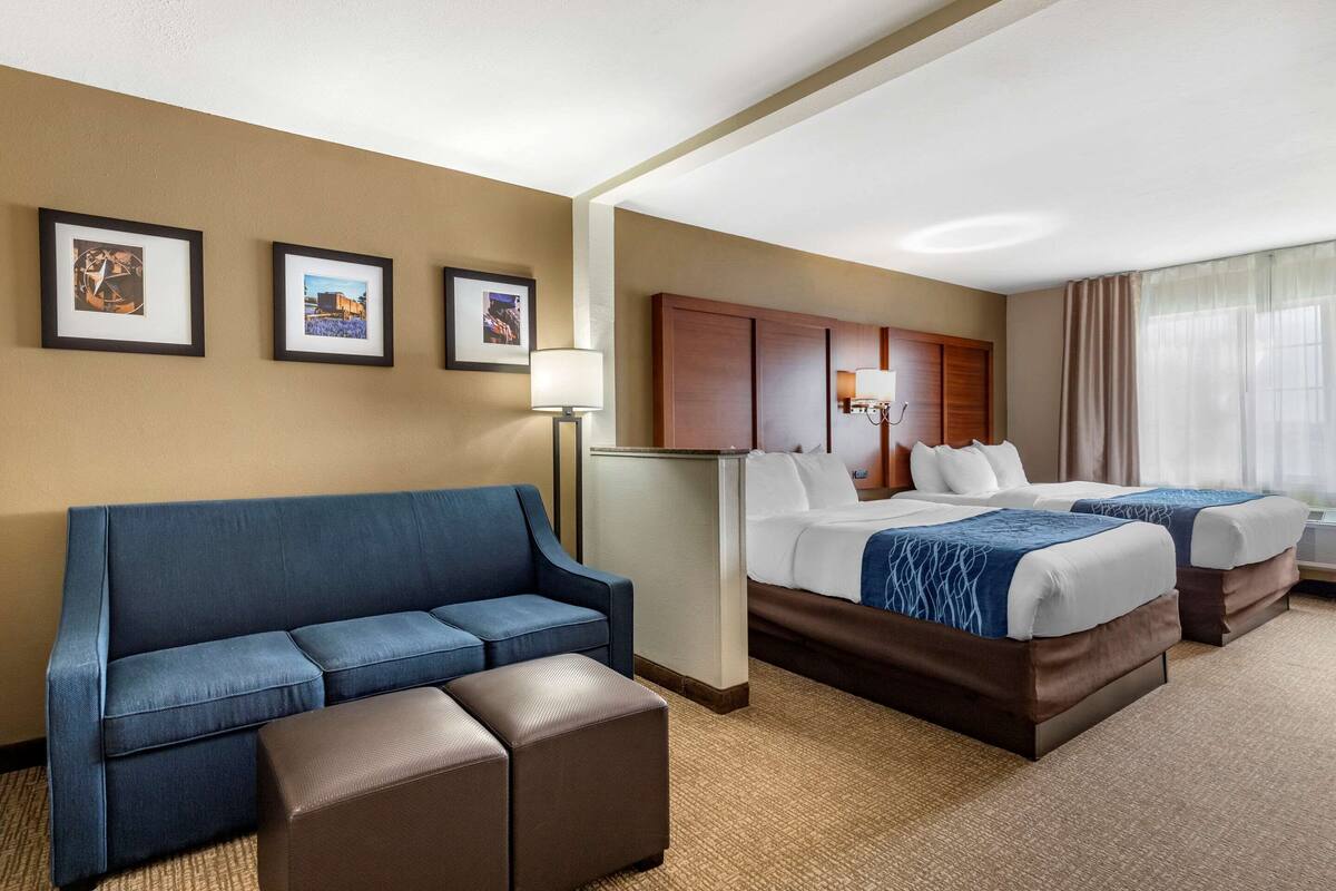 Habitación de hotel con un sofá azul, dos camas con ropa de cama blanca y azul, un cabecero de madera y cuadros enmarcados en la pared.