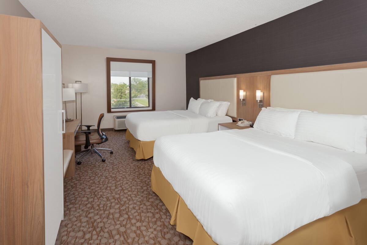 A hotel room with two beds, a desk with a chair, a lamp, and a window with a view.