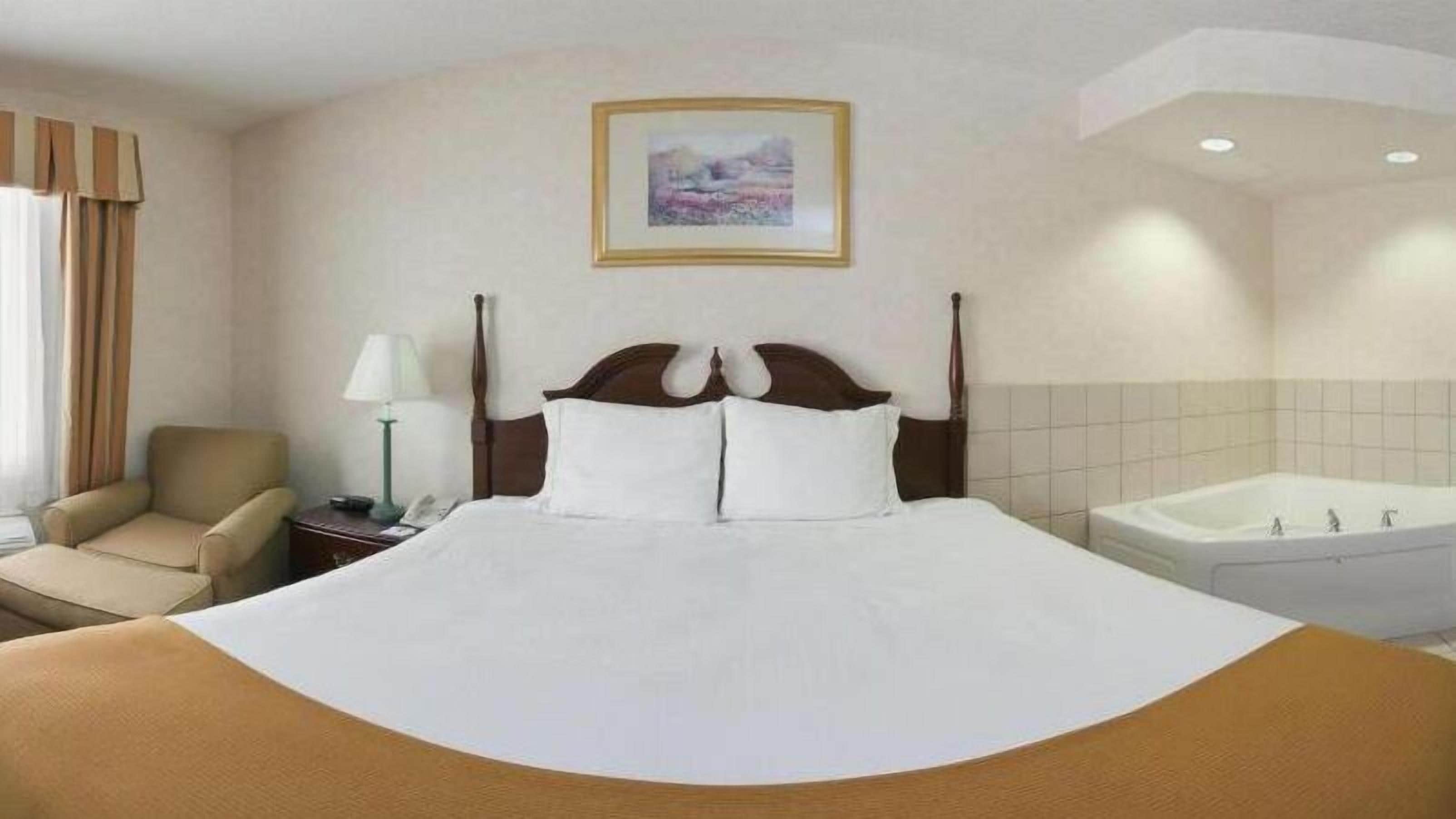 A hotel room with a large bed, a chair, a bedside table with a lamp, a framed picture on the wall, and a bathtub.