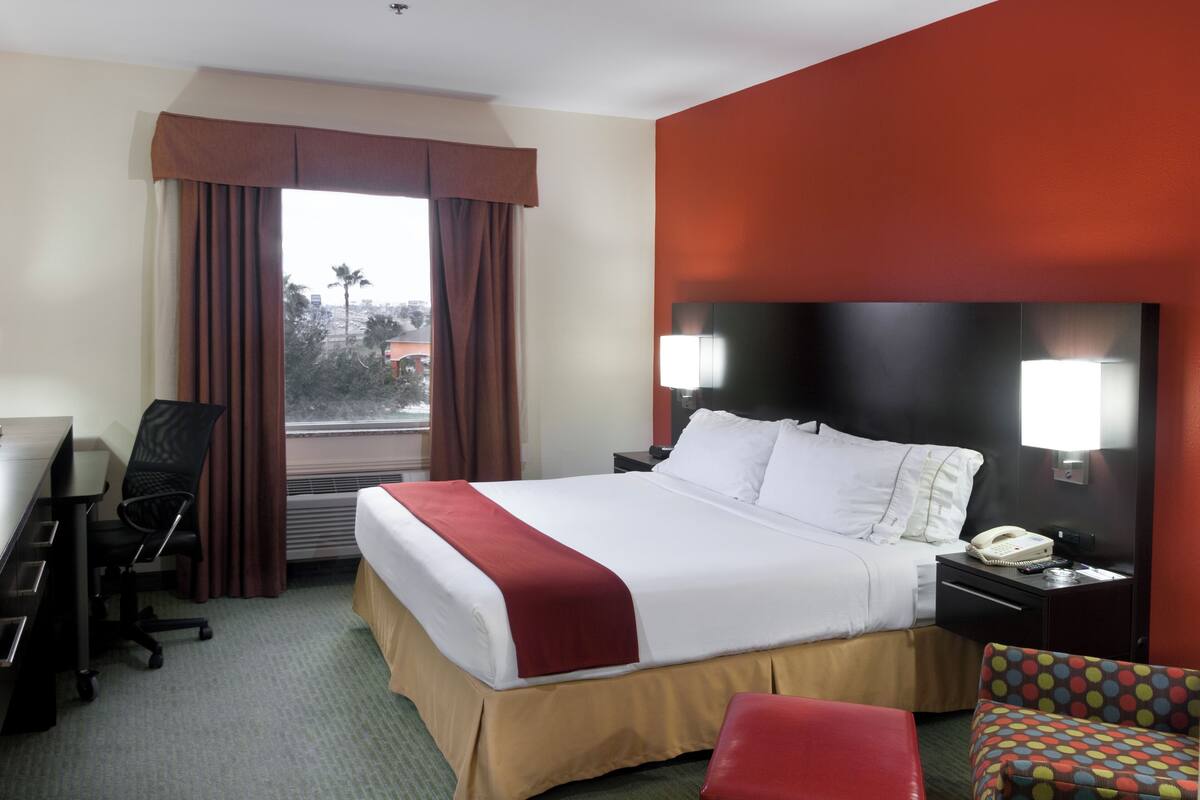 Una habitación de hotel con una cama grande, un escritorio, una silla, un otomano colorido y un ventanal con cortinas.