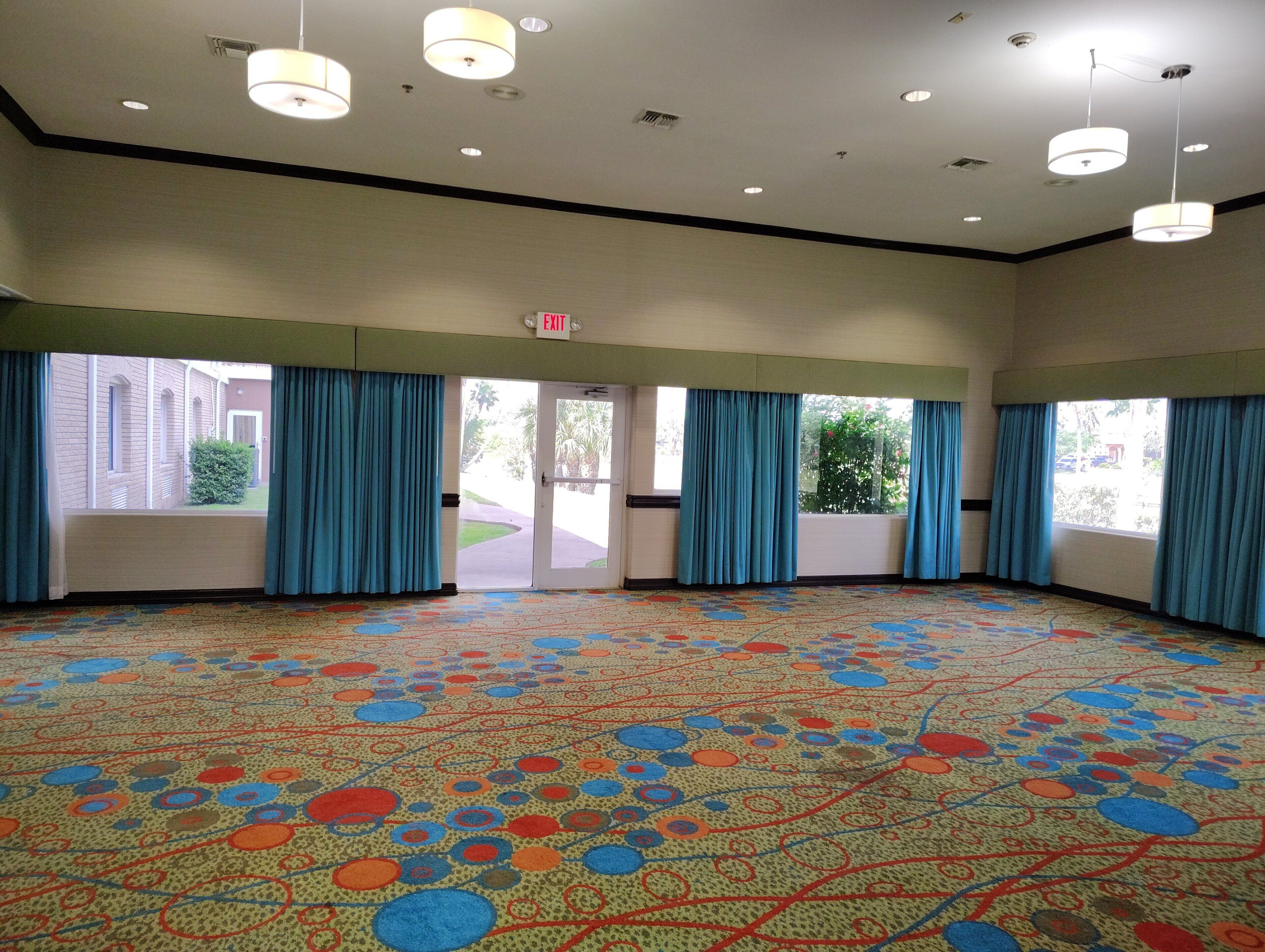 Amplia habitación con alfombra estampada, ventanas grandes con cortinas azules y múltiples luces de techo.