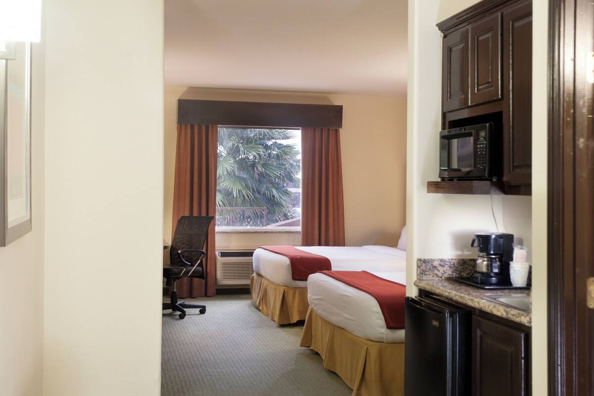 A hotel room with two beds, a kitchenette, a chair, and a window with curtains.