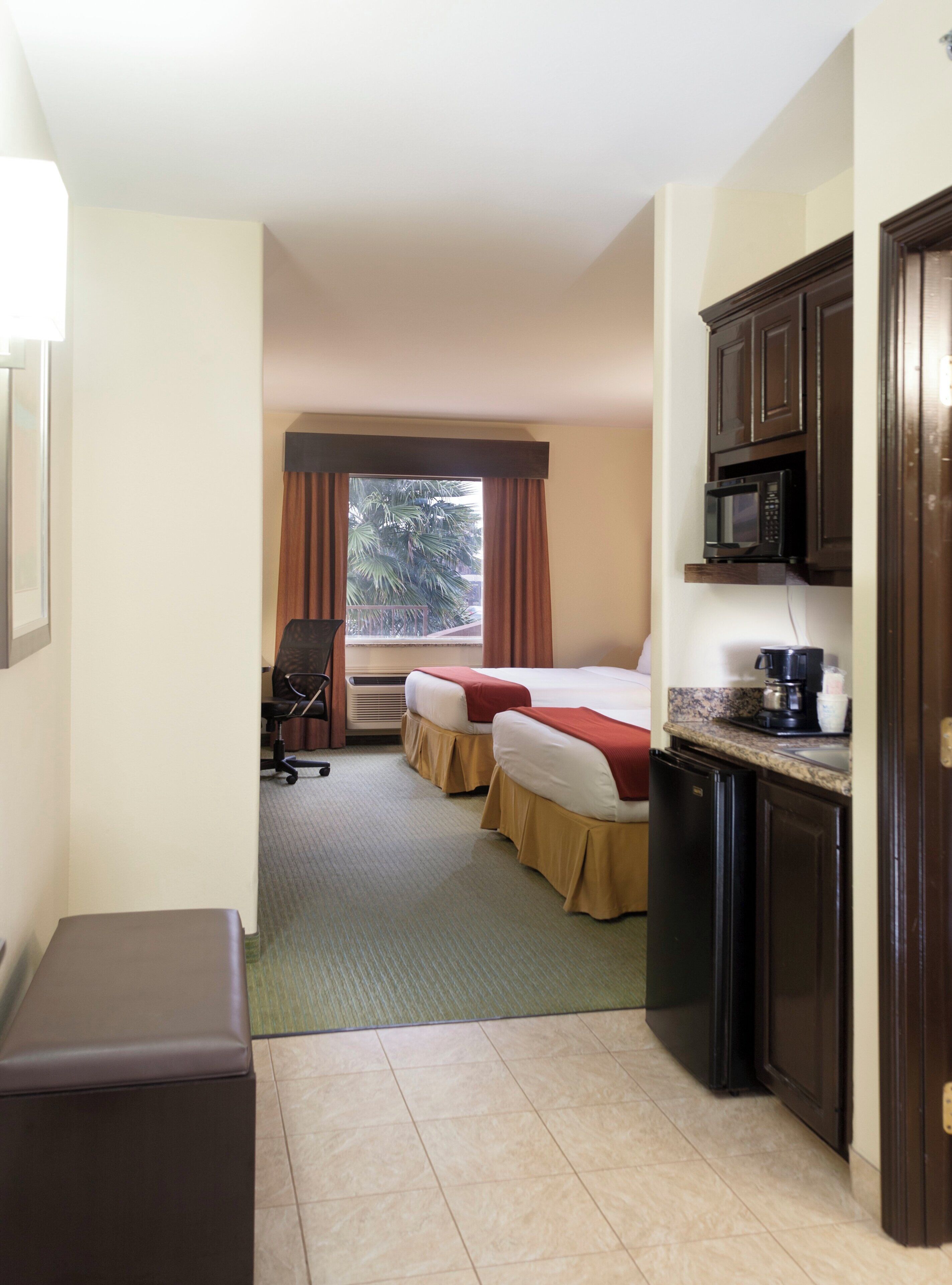 A hotel room with a kitchenette, a bed, a chair, and a window with curtains.
