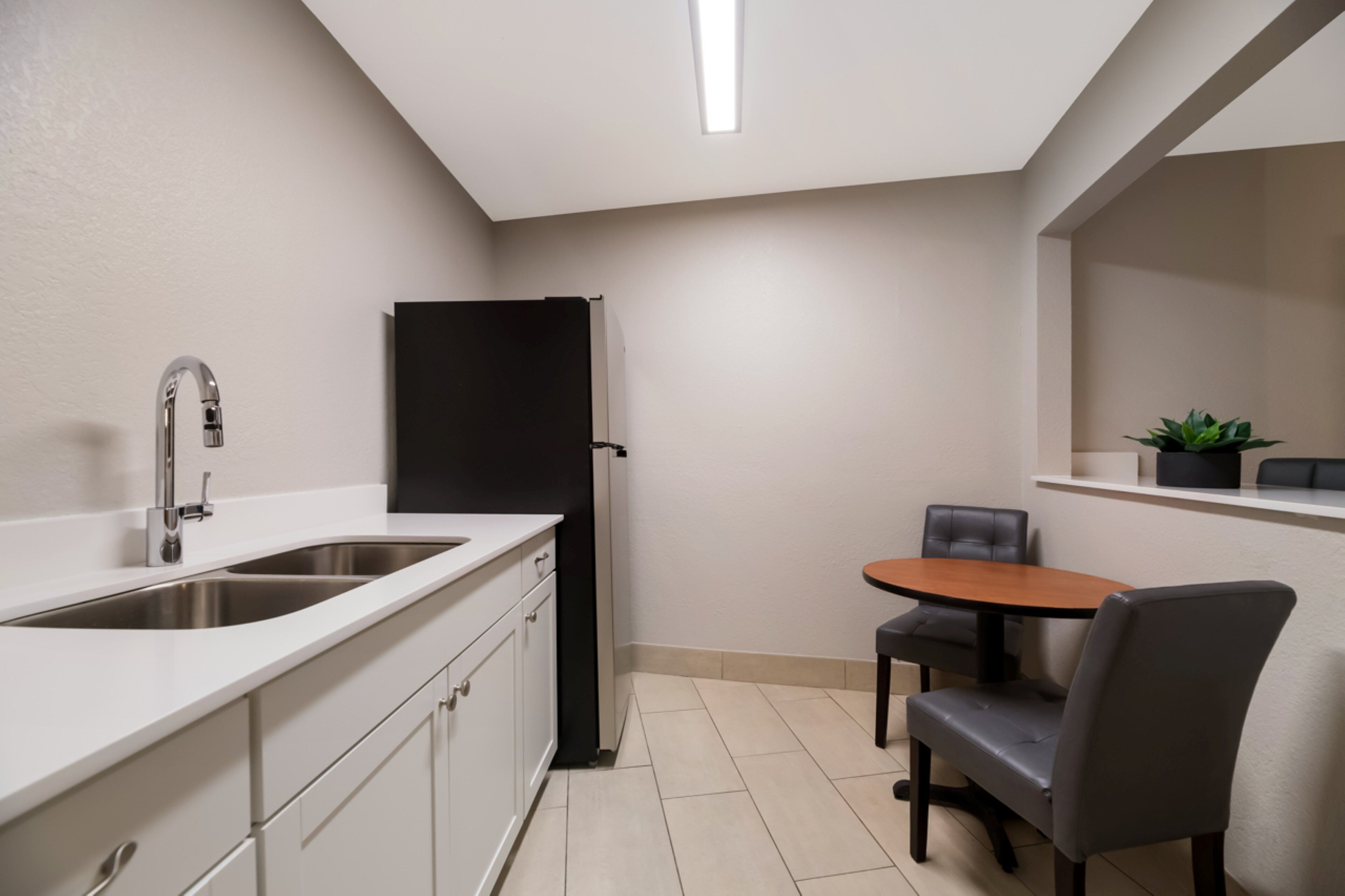 Cocina compacta con lavaplatos, un refrigerador negro, una mesa de comedor con dos sillas y una planta en maceta sobre una repisa.