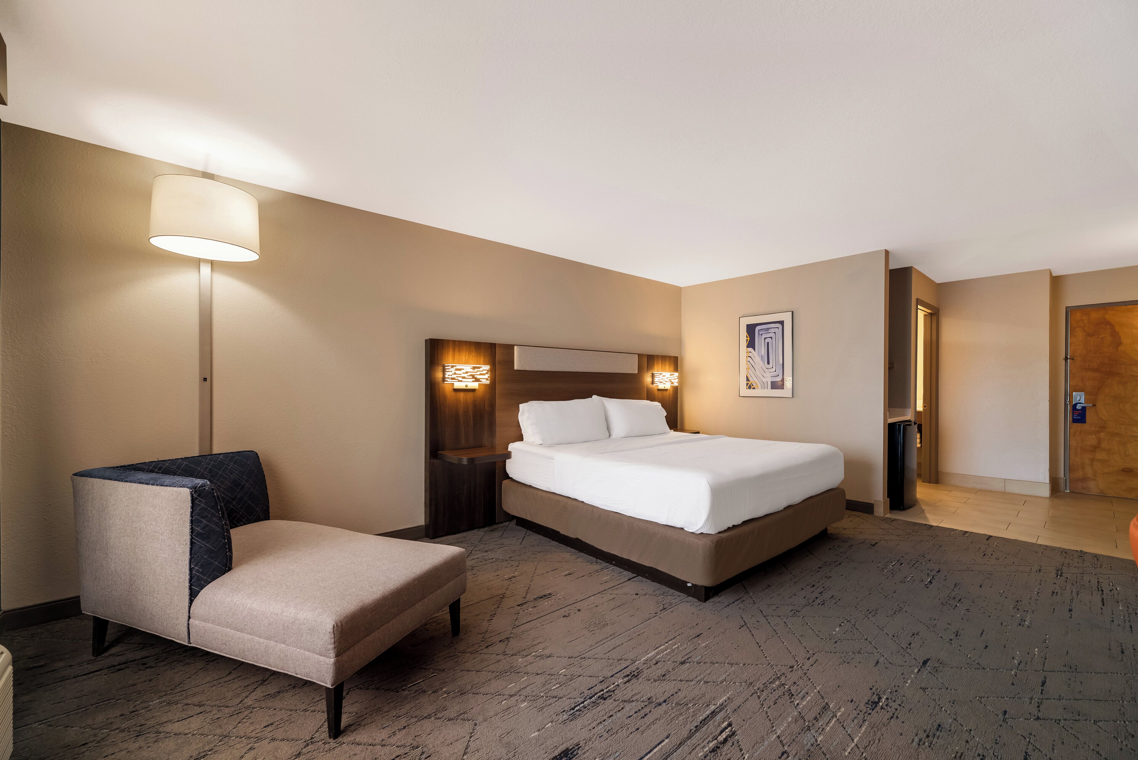 Une chambre d’hôtel avec un lit, une chaise, une table de chevet, une lampe et un tableau au mur.