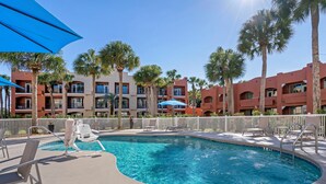 A swimming pool with lounge chairs and umbrellas, surrounded by palm trees and a multi-story building with balconies. - Holiday Inn Express Hotel & Suites The Villages by IHG (Lady Lake)