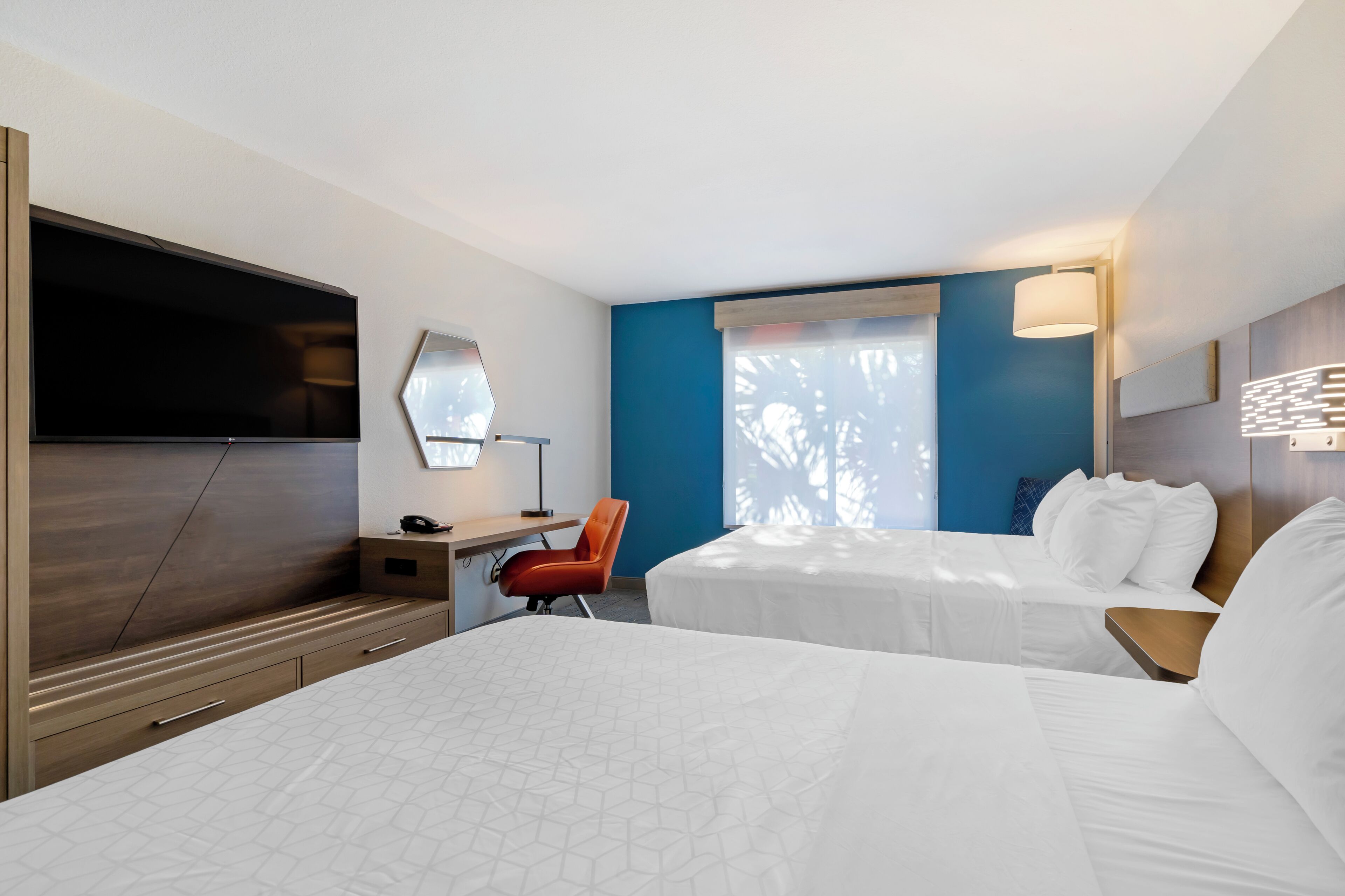 Quarto de hotel com duas camas, uma escrivaninha com cadeira, televisão e janela com persianas.
