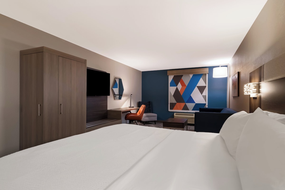 A hotel room with a large bed, a desk with a chair, a wardrobe, and a colorful geometric wall art.