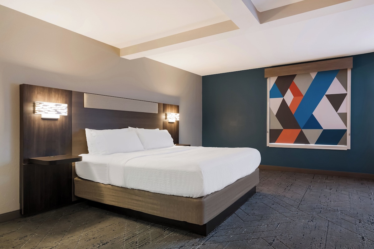 A modern hotel room with a large bed, wooden headboard, and a geometric wall art piece.