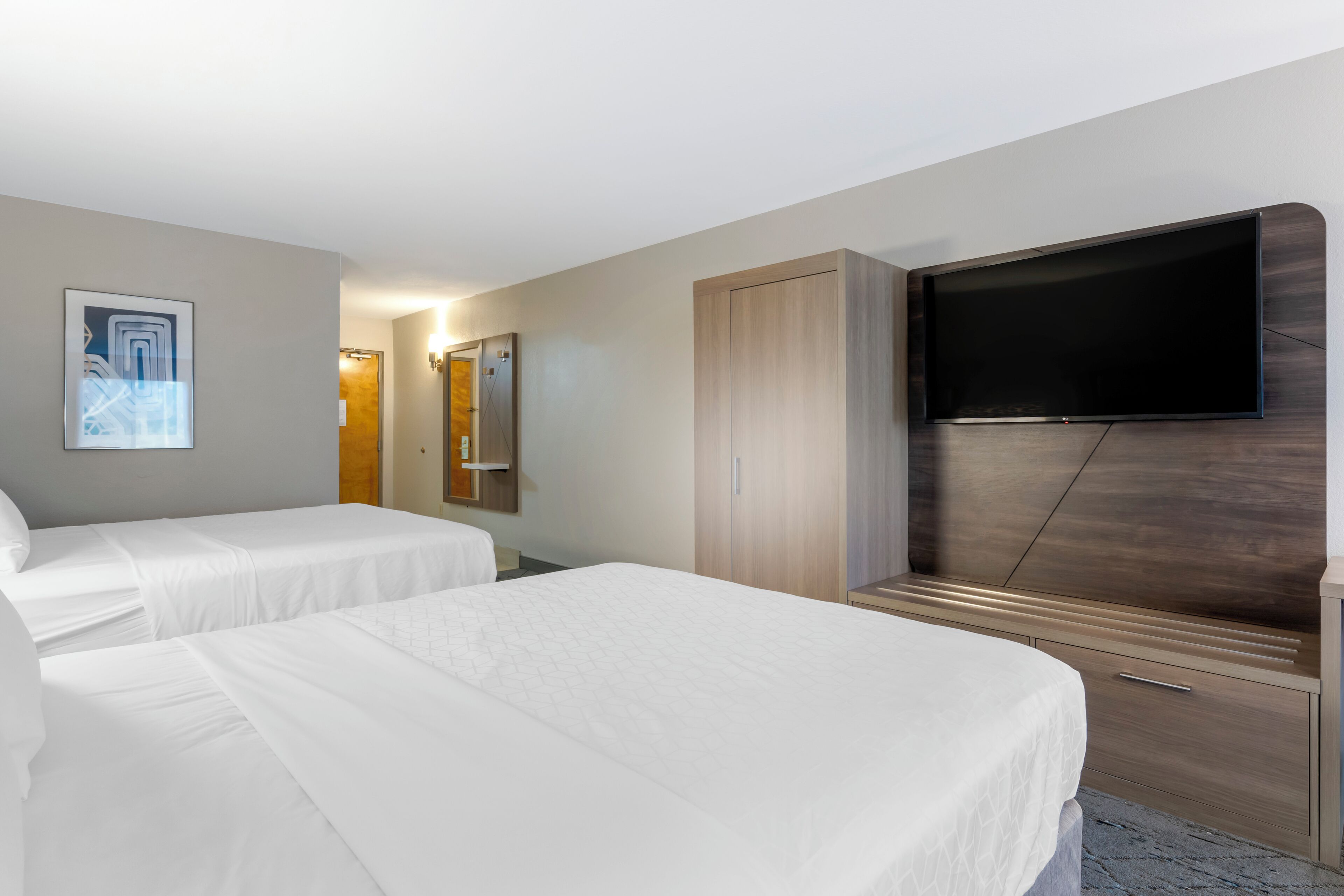 A hotel room with two beds, a flat-screen TV mounted on a wooden panel, a wardrobe, and a mirror.