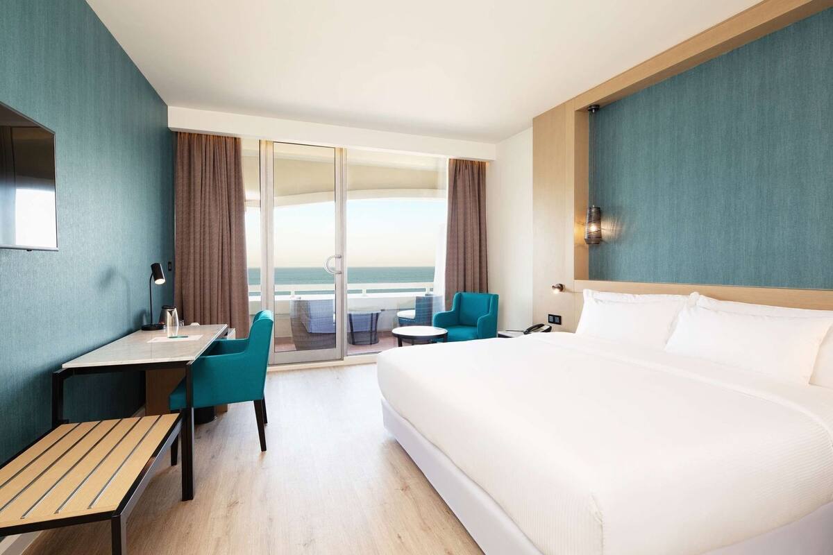 A hotel room with a large bed, a desk with a chair, a small table, and a view of the ocean through a sliding door.