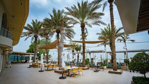 An outdoor seating area with tables and chairs, surrounded by palm trees, under a shaded canopy. - Occidental Sharjah Grand (Sharjah)