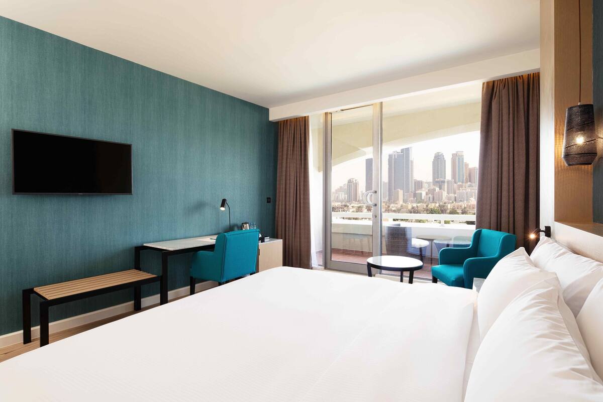 A hotel room with a large bed, a desk, a chair, and a view of a cityscape through a balcony.