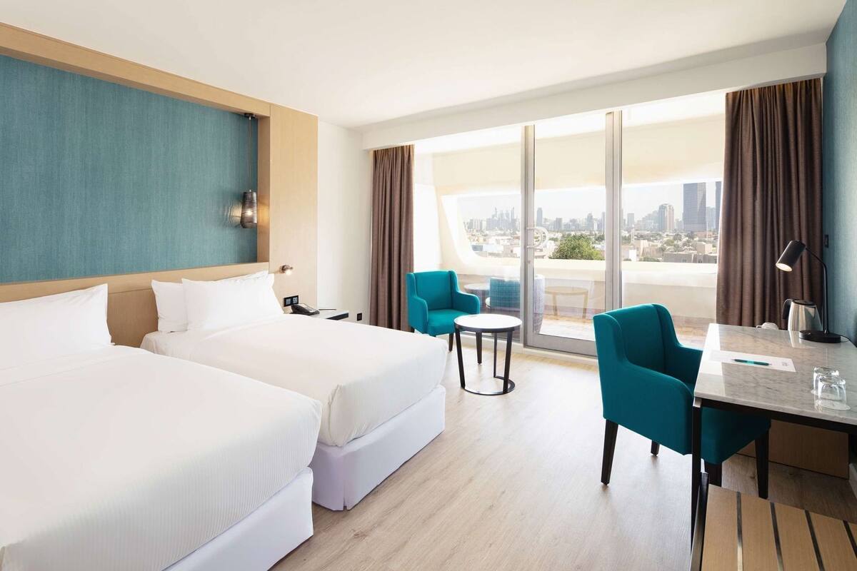A hotel room with two beds, a balcony with a city view, a small table with chairs, and a desk with a lamp.