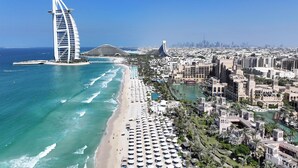 A coastal cityscape with a prominent sail-shaped hotel, a beach with umbrellas, and a marina. - Occidental Sharjah Grand (Sharjah)