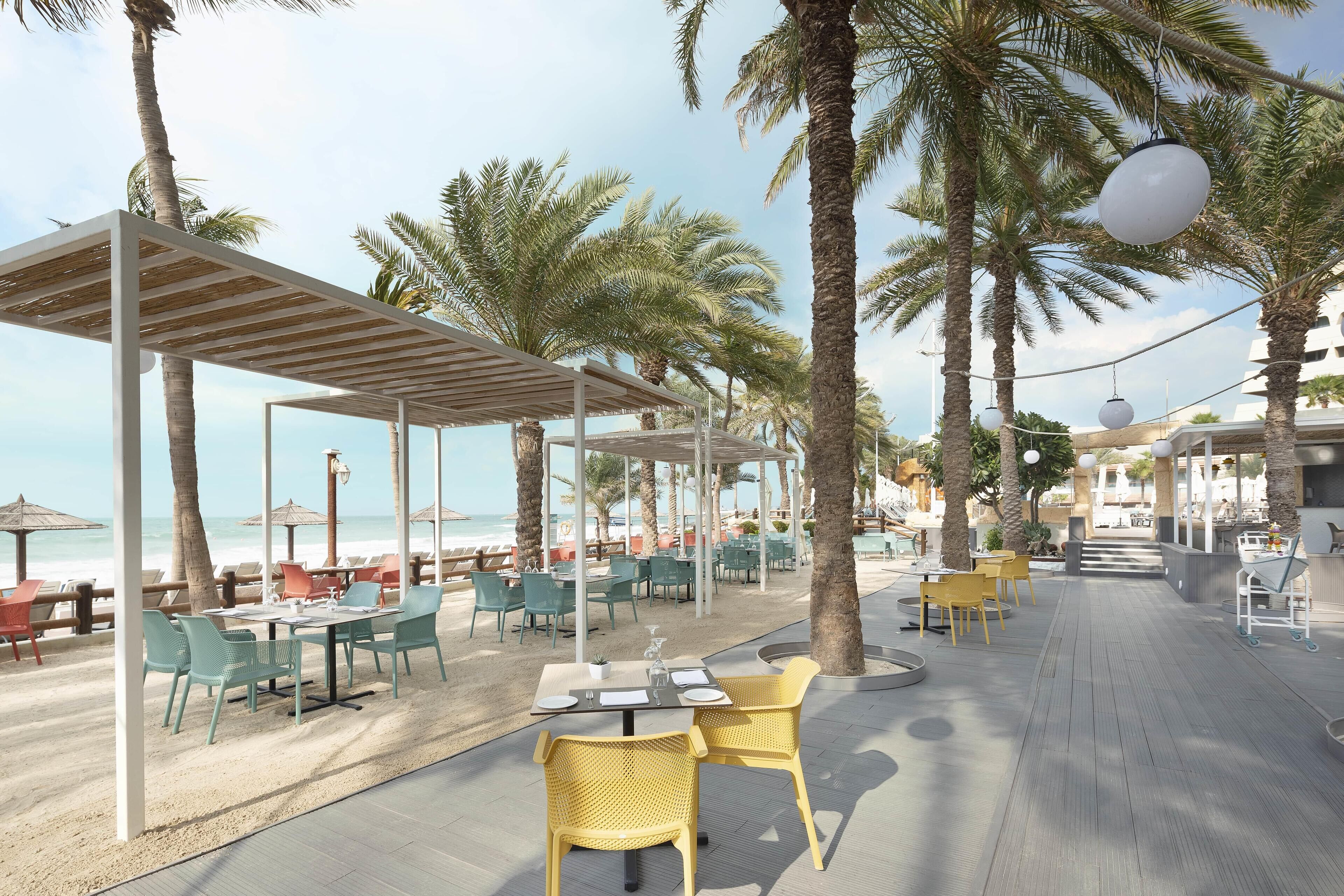 An outdoor dining area with tables and chairs, palm trees, and a view of the beach.