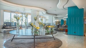 A modern hotel lobby with a round table surrounded by vases of yellow flowers, a teal accent wall, and a glass railing overlooking the city. - Occidental Sharjah Grand (Sharjah)
