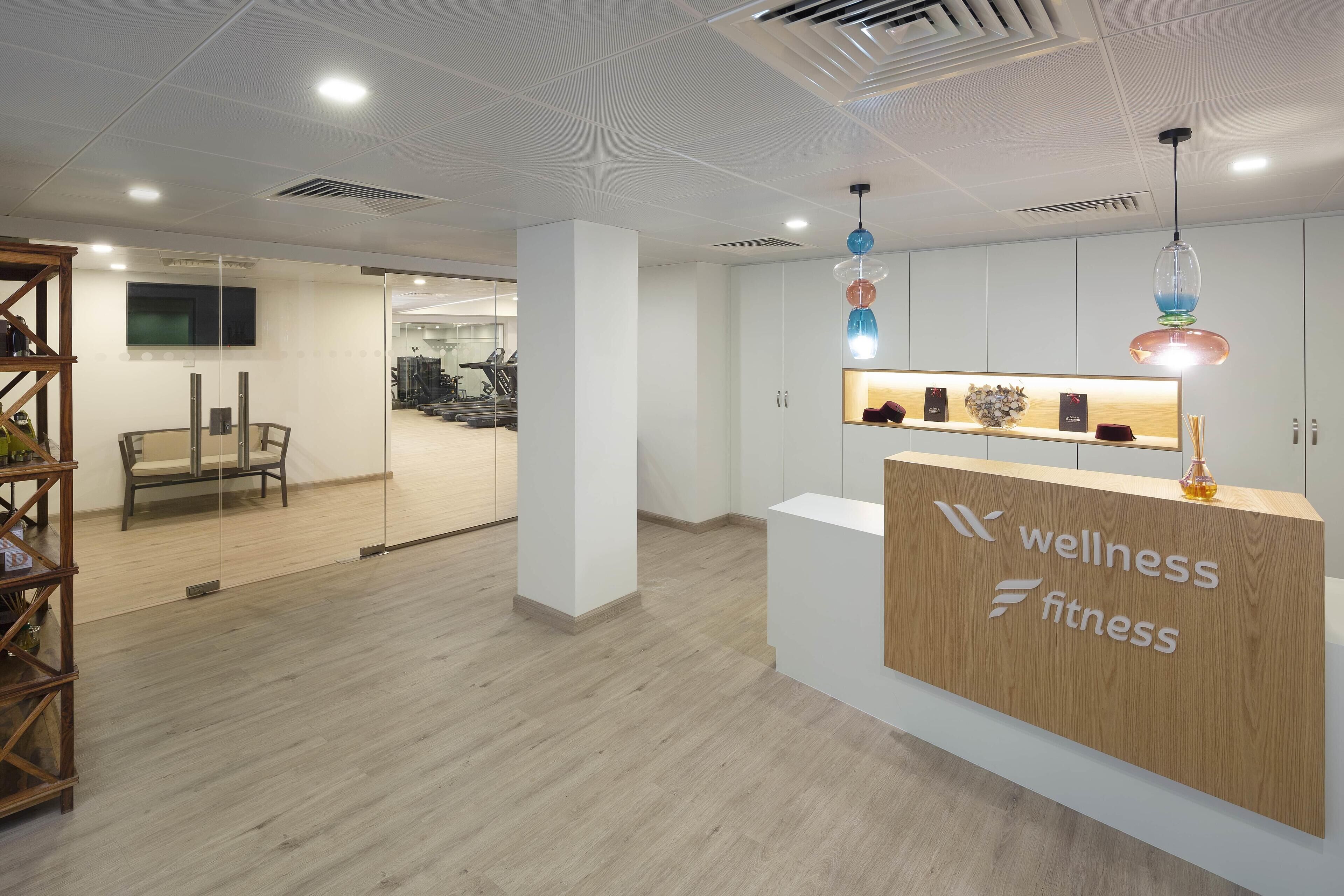 Zona de recepción con un mostrador de madera identificado como "wellness fitness", una pared de vidrio y un área de ejercicios visible al fondo.