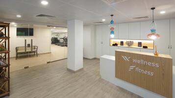 Zona de recepción con un mostrador de madera identificado como "wellness fitness", una pared de vidrio y un área de ejercicios visible al fondo.